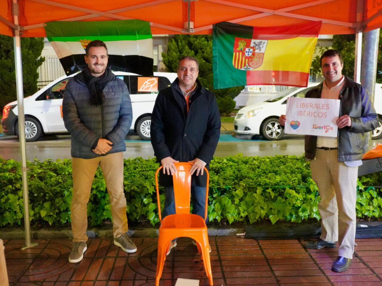 Tres hombres en un evento político con banderas ibéricas y un cartel