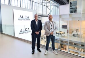 Dos personas de pie en DFactory Barcelona junto al logo de Alca Technologies.