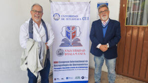 Dos hombres posando frente a un cartel del congreso de antropología en México.