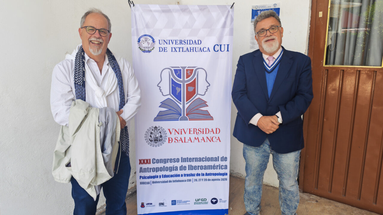 Dos hombres posando frente a un cartel del congreso de antropología en México.
