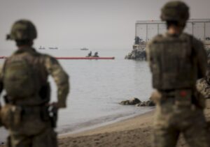 Soldados observando una operación en la costa con barcos en el agua.