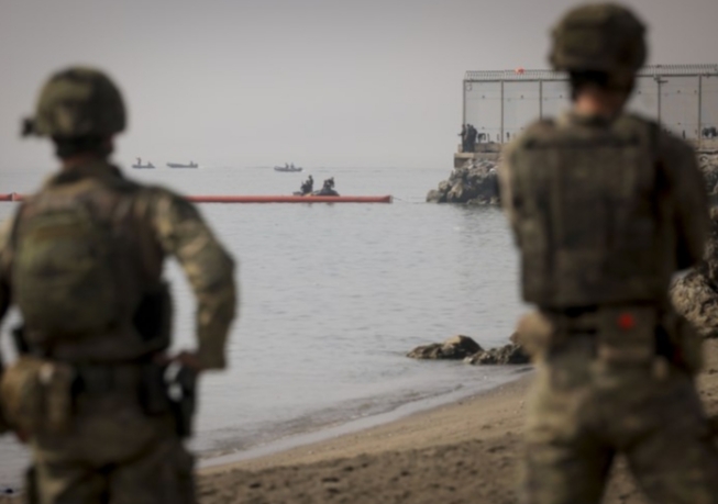 Soldados observando una operación en la costa con barcos en el agua.