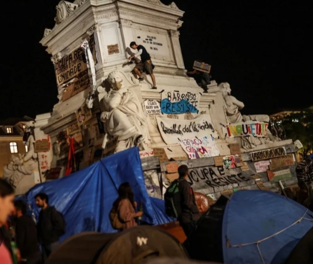 Personas y tiendas de campaña rodean un monumento cubierto de pancartas durante la noche