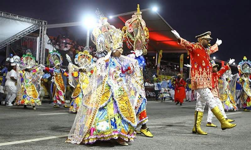 Comparsa de carnaval desfila con trajes brillantes frente a gradas iluminadas.