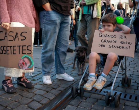 Manifestantes en una calle sostienen carteles sobre vivienda, con una persona sentada en primer plano.