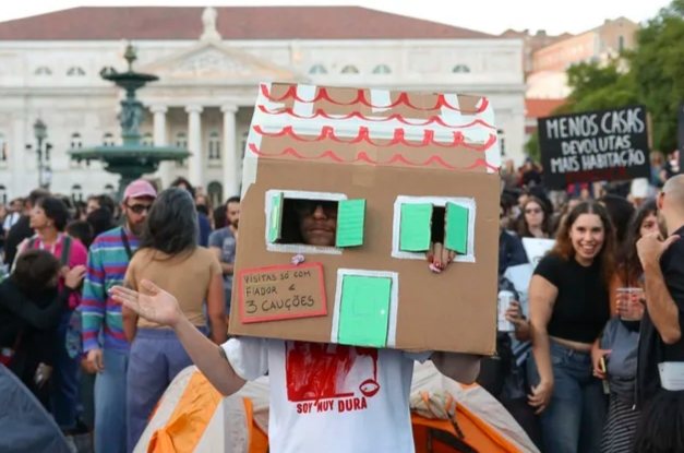 Manifestante con una casa de cartón entre una multitud reunida en una plaza.