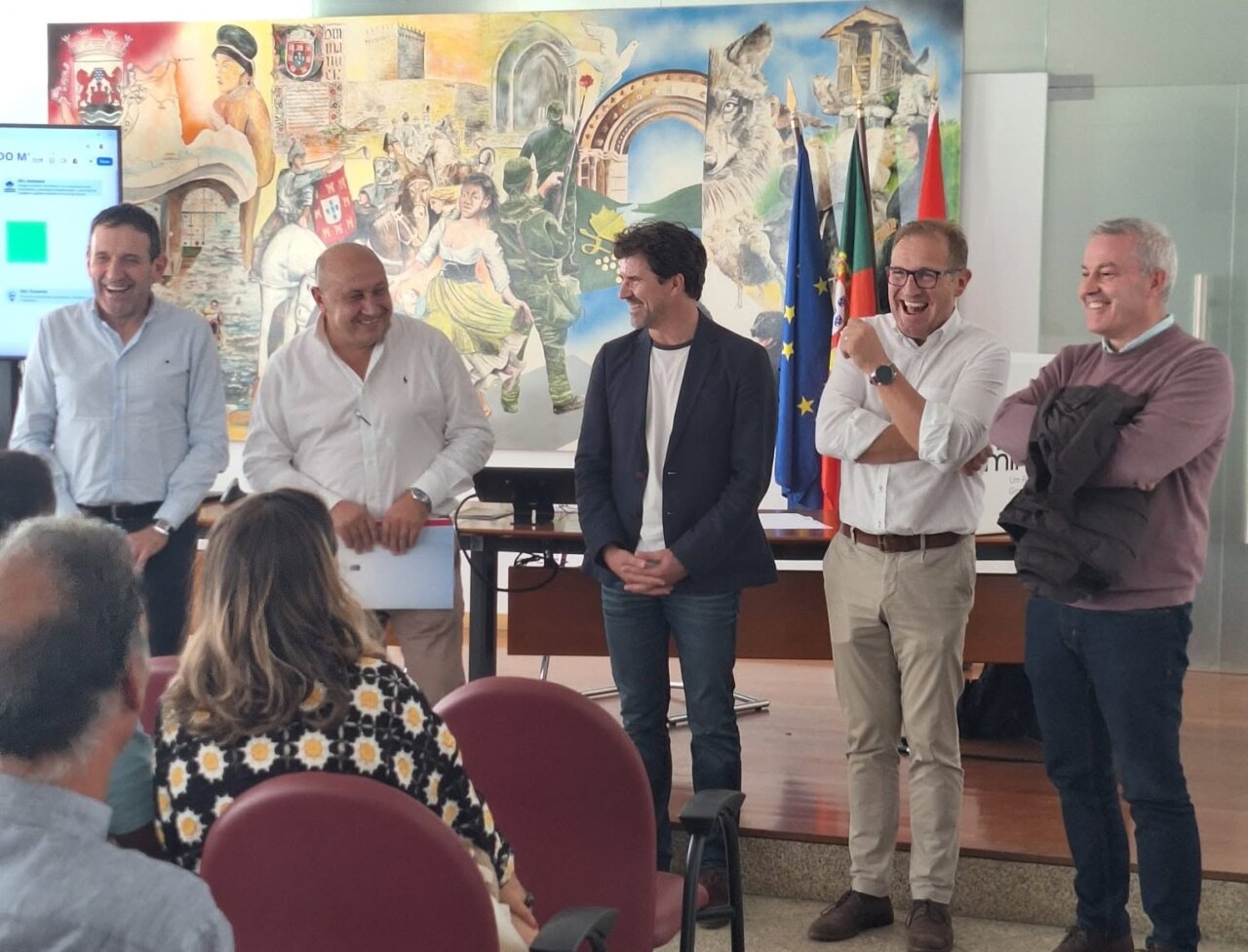 Varias personas conversan frente a un mural, con asistentes sentados y banderas al fondo